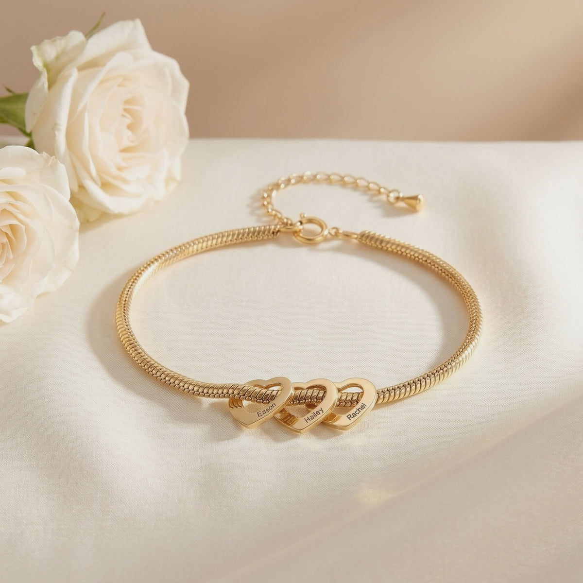 Gold-tone snake chain bracelet with three engraved heart charms displayed flat on white satin beside white roses.