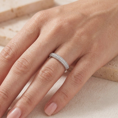 Silver triple-row micro-pavé ring with clear stones worn on a person's hand over a beige textured surface.