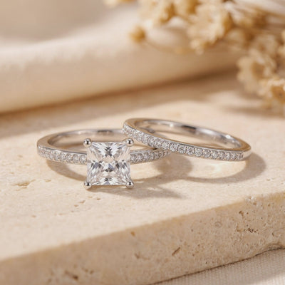 Silver ring set displayed on a stone surface, showing the princess-cut center stone and matching pavé band.