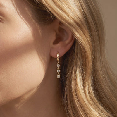 Gold huggie hoop drop earring with three round bezel-set stones worn on a woman's ear in close-up.