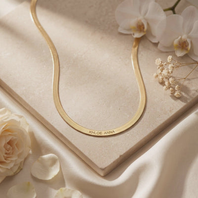 Gold-toned flat herringbone chain necklace with KHLOE ANNA engraving, laid on a stone tile beside white flowers.