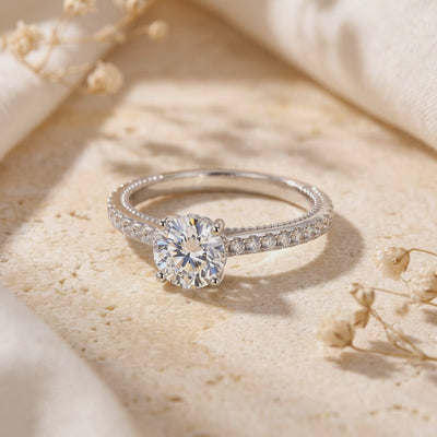 Silver ring with a round clear center stone and four-prong setting, resting on a beige textured surface.
