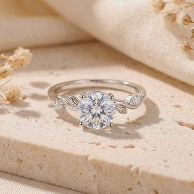 Close-up of a silver leaf-motif ring with a round clear center stone resting on a beige stone slab.