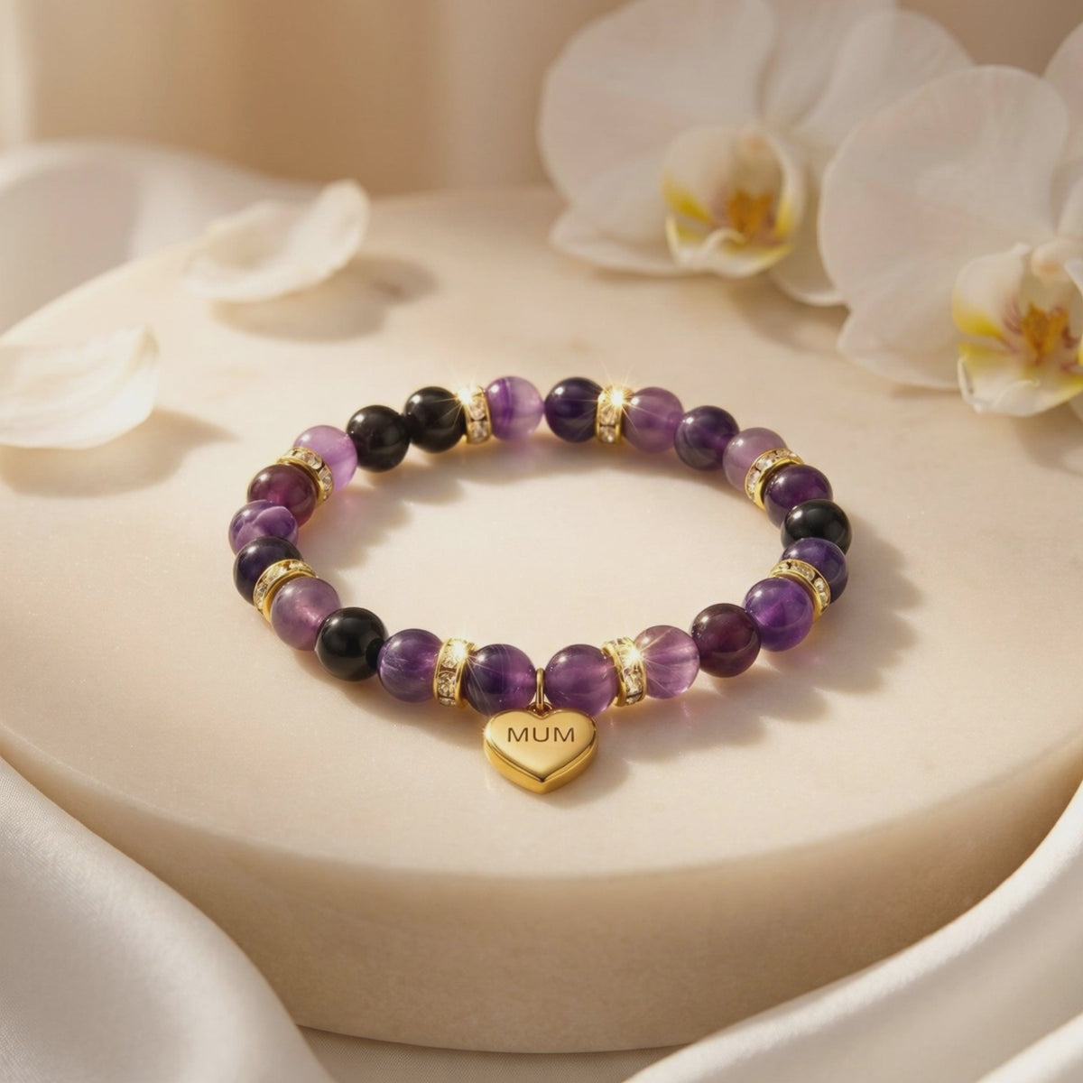 Purple amethyst and black beaded bracelet with a gold-tone “MUM” heart charm displayed on a round white pedestal