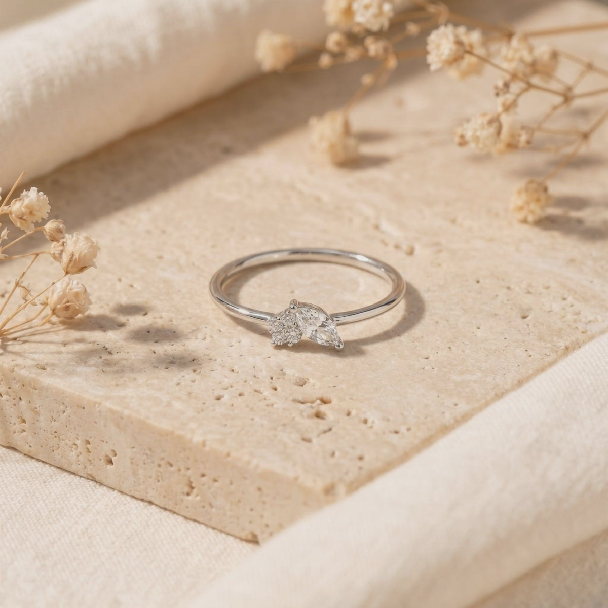 Silver marquise moissanite cluster ring resting flat on a textured stone slab alongside dried flowers.