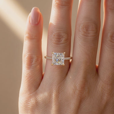 A yellow gold-toned solitaire ring with a princess-cut moissanite and pavé band, worn on a finger.