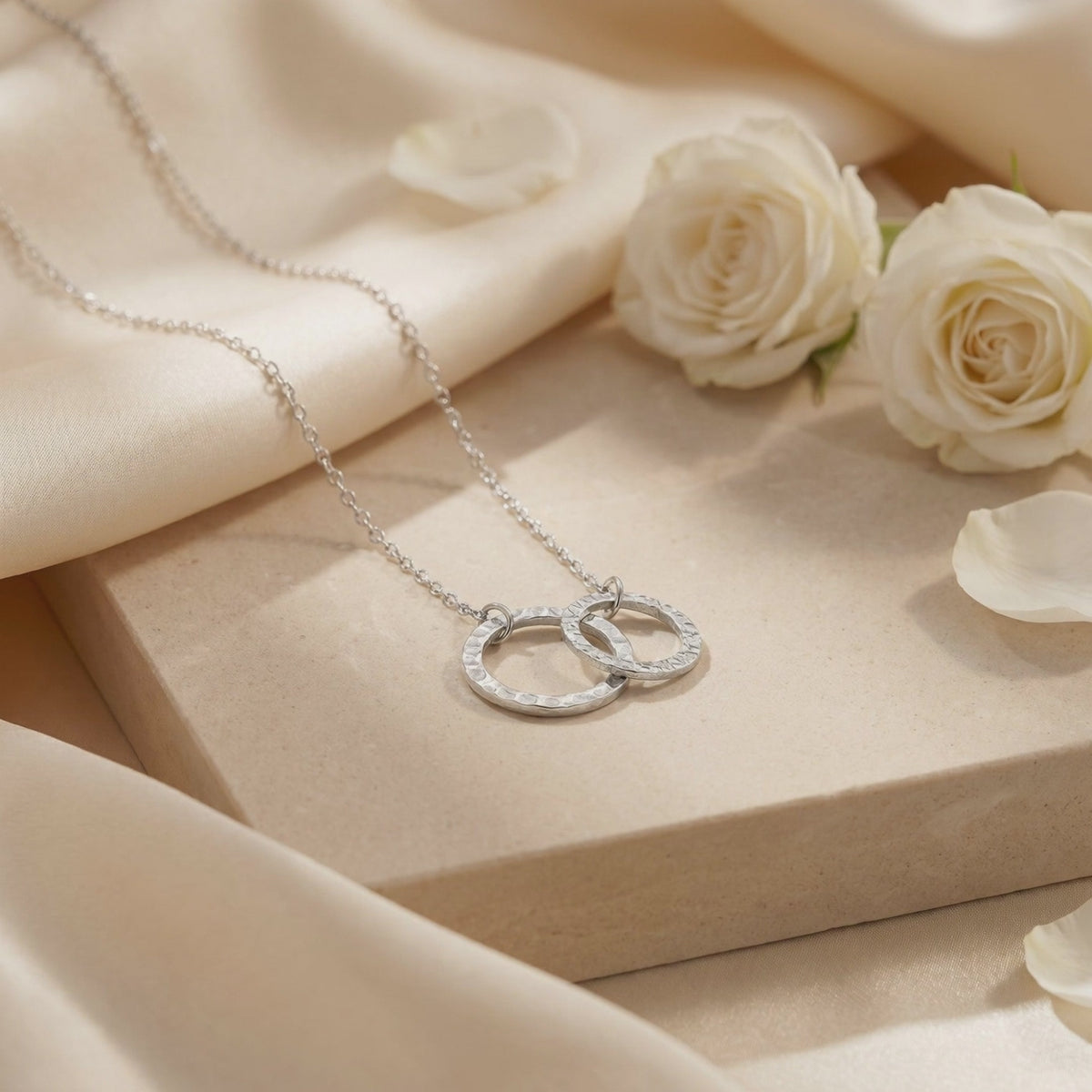 Silver-tone necklace with textured interlocking circles arranged on a beige display block beside white roses