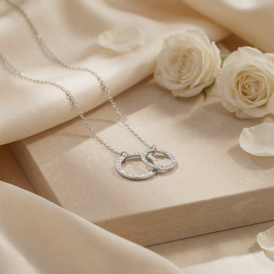 Silver-tone necklace with textured interlocking circles arranged on a beige display block beside white roses