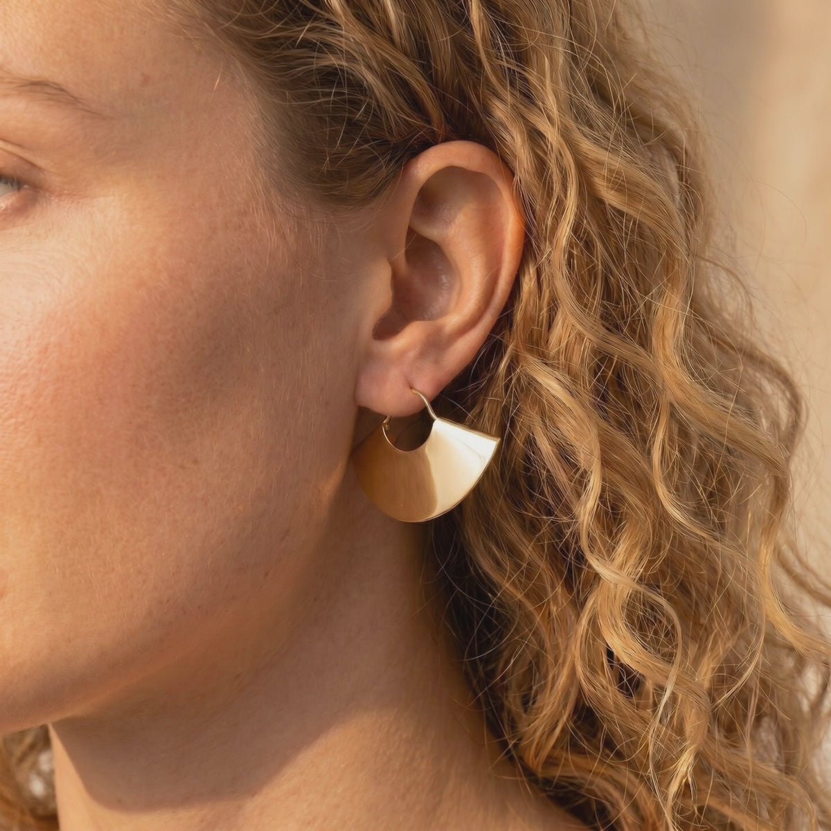 Close-up side profile of a woman wearing a gold-tone fan-shaped drop earring with curly hair visible.