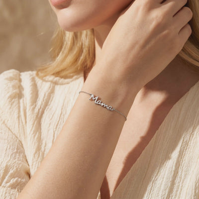 Silver-toned Mama script chain bracelet worn on a woman's wrist over cream-colored fabric.