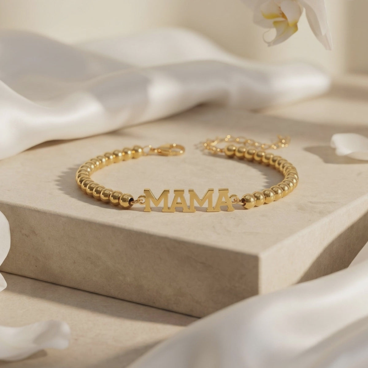 Gold-tone beaded MAMA bracelet displayed on a beige platform with its clasp and extender chain visible.