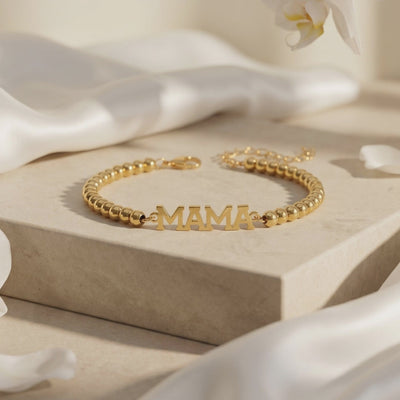 Gold-tone beaded MAMA bracelet displayed on a beige platform with its clasp and extender chain visible.