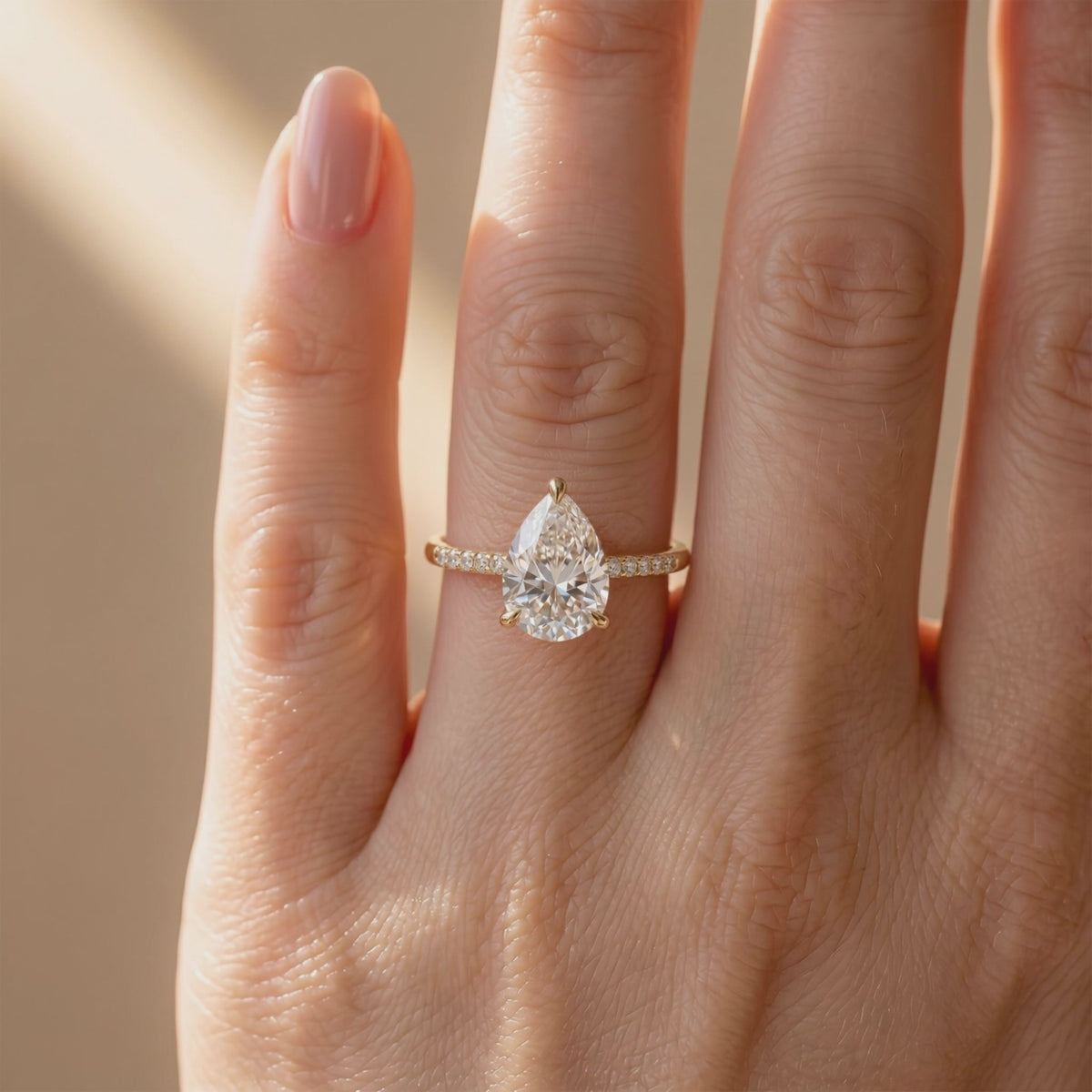 Gold-tone pear-cut moissanite solitaire ring with a pavé band, shown worn on a finger.