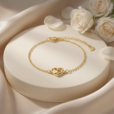 Gold-tone chain bracelet with a heart-shaped cutout charm on a cream display stand beside white roses.