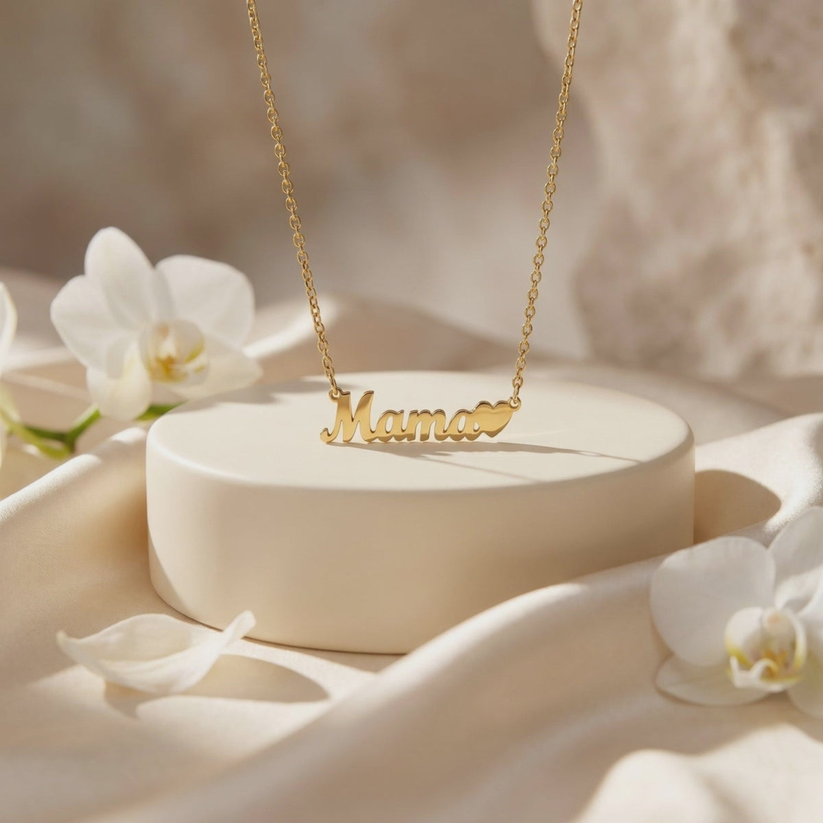 Gold-tone Mama script necklace with a heart detail displayed on a cream round podium beside white orchids.