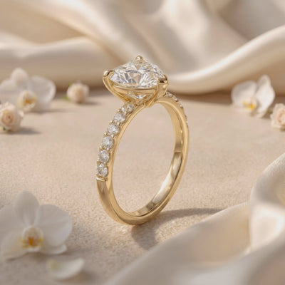 Angled side view of a heart-cut solitaire ring with a pavé band on cream fabric among white flowers.
