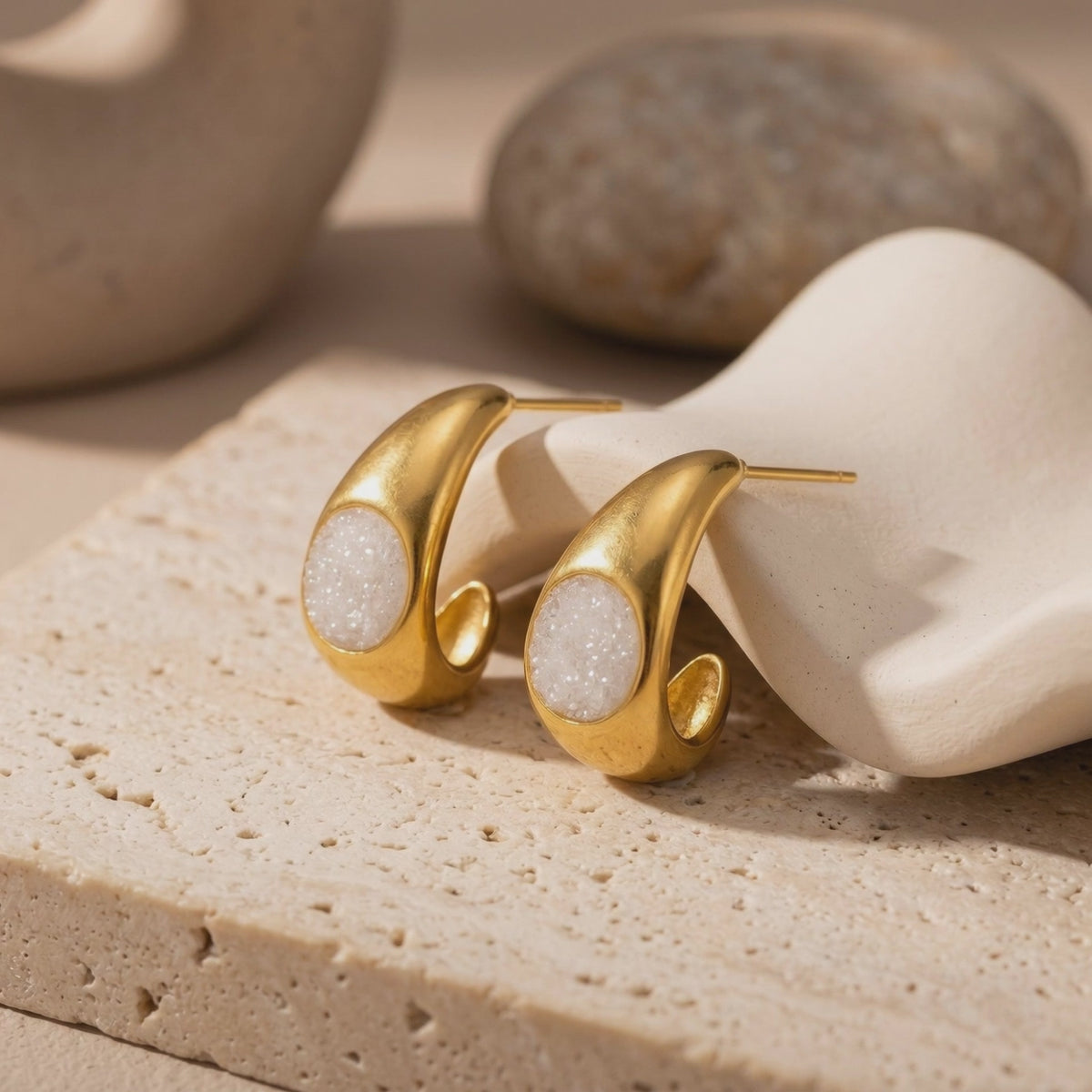 Pair of gold teardrop earrings with white textured insets displayed on light-colored textured blocks.