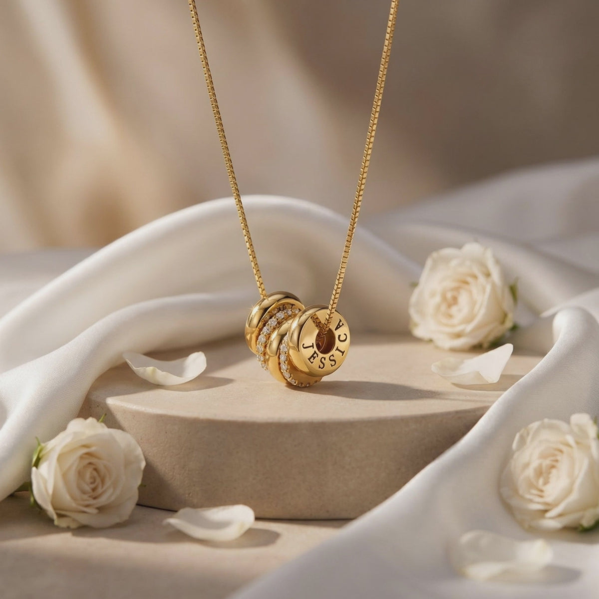 Gold-tone engraved ring charm necklace displayed upright on a beige pedestal with white roses and draped fabric.