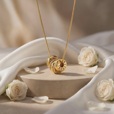 Gold-tone engraved ring charm necklace displayed upright on a beige pedestal with white roses and draped fabric.