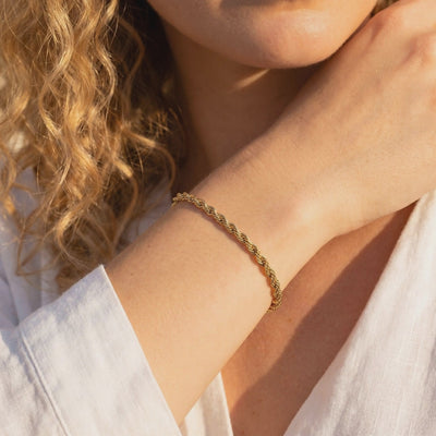 Gold-toned rope bracelet worn on a wrist in warm sunlight, with a white sleeve visible.