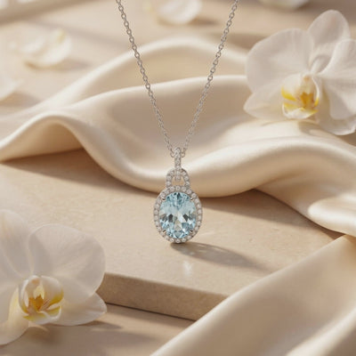 Silver necklace with an oval light-blue simulated aquamarine pendant displayed on cream fabric among white orchids
