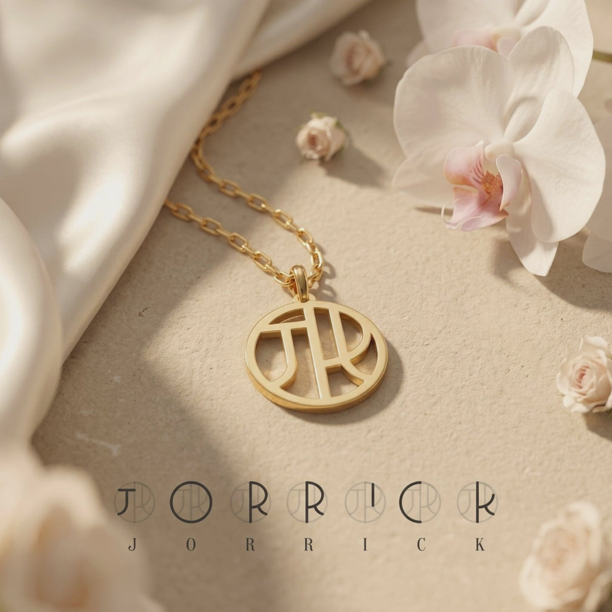 Gold-toned circular monogram necklace displayed flat with white orchids and light-colored draped fabric