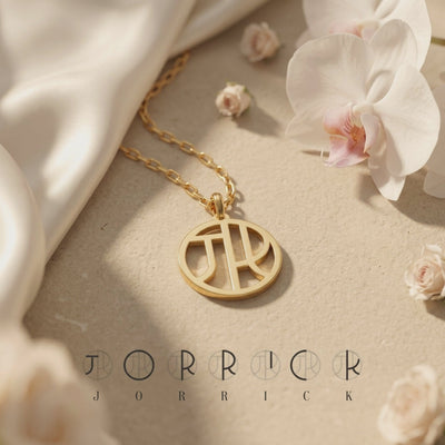 Gold-toned circular monogram necklace displayed flat with white orchids and light-colored draped fabric