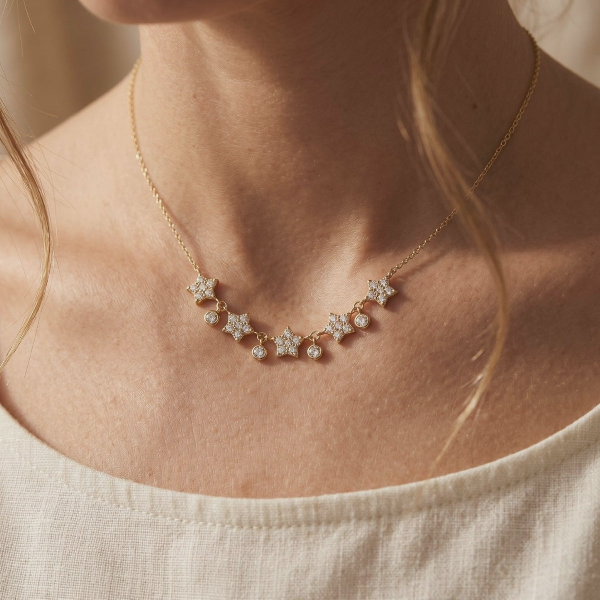 Gold-tone necklace worn around a model's collarbone, featuring five jeweled stars and four dangling round bezels.