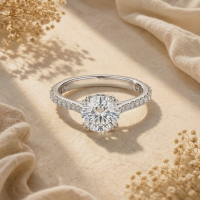 Silver-toned round moissanite ring with pavé shoulders displayed on beige fabric beneath floral shadows.