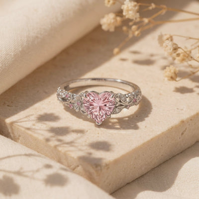 Close-up view of The Rosalique Ring, a solid gold ring with a heart-shaped pink simulated diamond centerpiece and floral filigree band, resting on a stone surface.