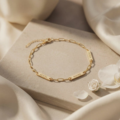 Gold-toned chain bracelet with three engraved name plates displayed flat on a stone slab.