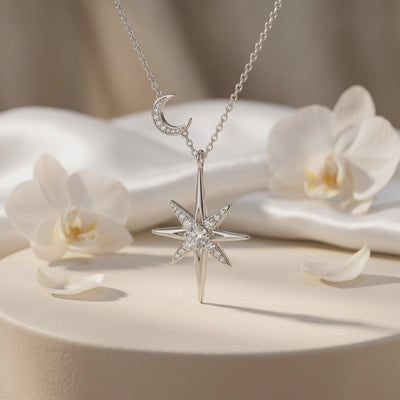Silver-tone starburst and crescent moon necklace displayed on a draped pedestal beside white orchids.