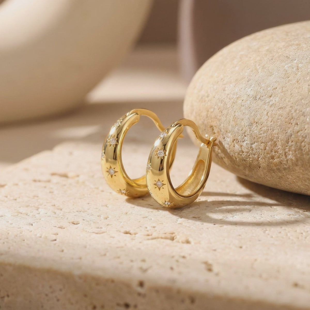 Pair of gold-tone starburst huggie hoop earrings standing on a pale textured stone surface beside a rounded stone.