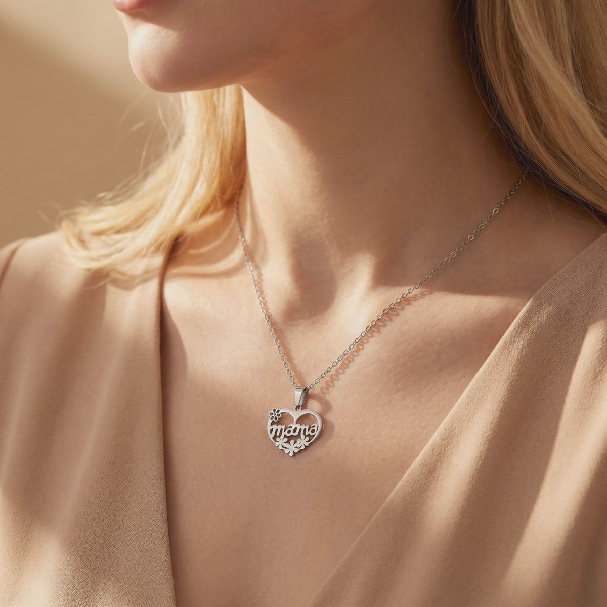 Silver heart necklace with mama script and floral cutouts worn around a model's neck over a beige top.