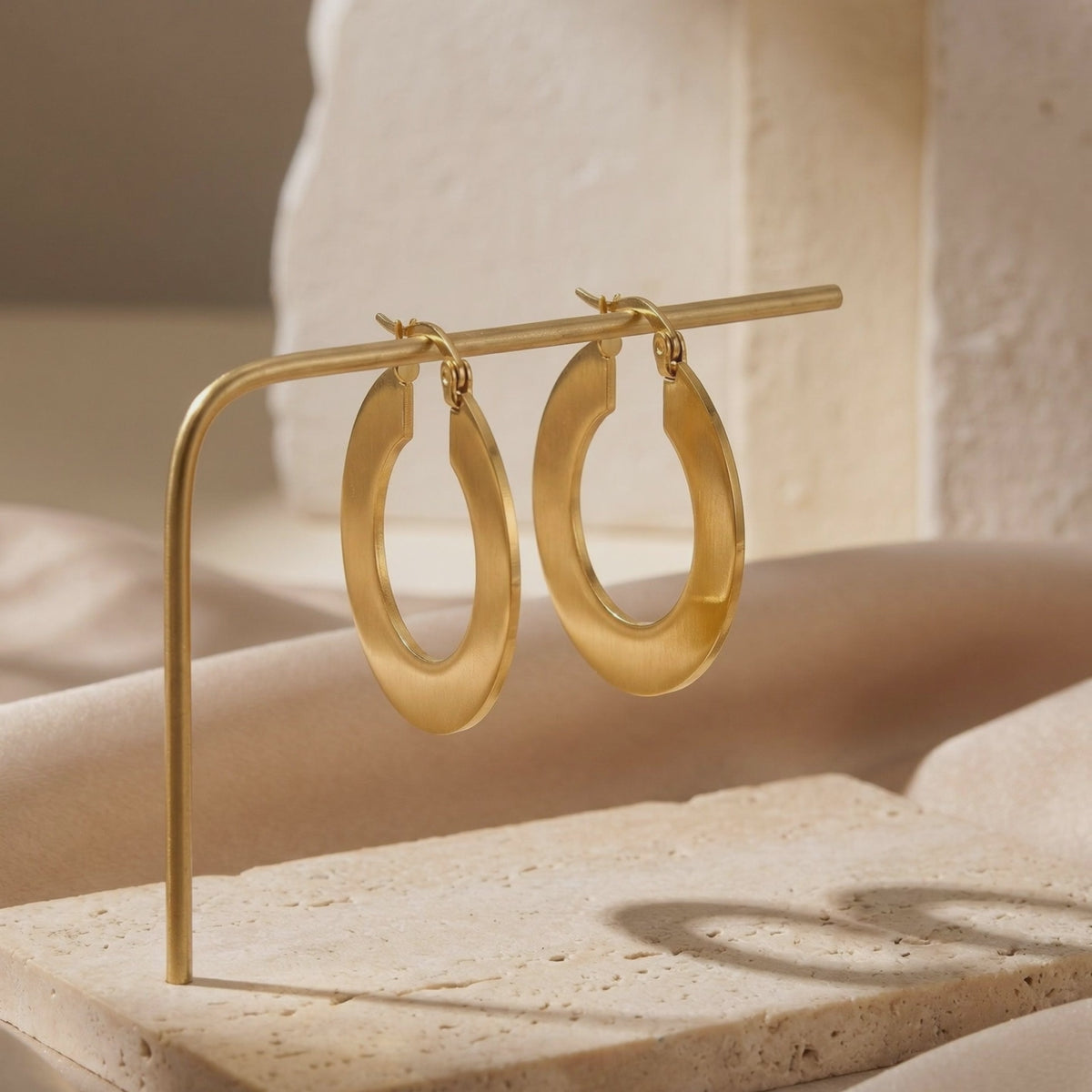 Pair of gold-tone wide hoop earrings hanging from a thin metal stand over a stone display surface.
