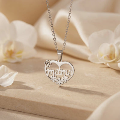 Close-up of a silver mama heart pendant with floral cutouts and chain, displayed on a beige surface with orchids.