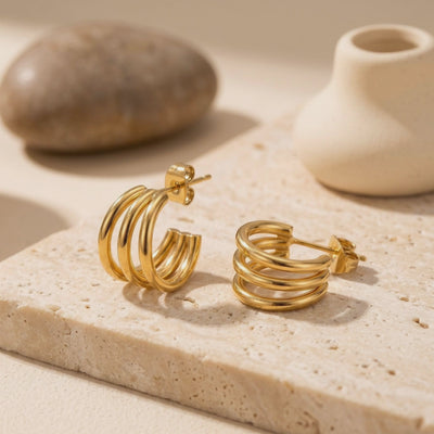 Pair of gold-tone triple-band hoop earrings displayed on a textured stone block with ceramic vessels behind them.