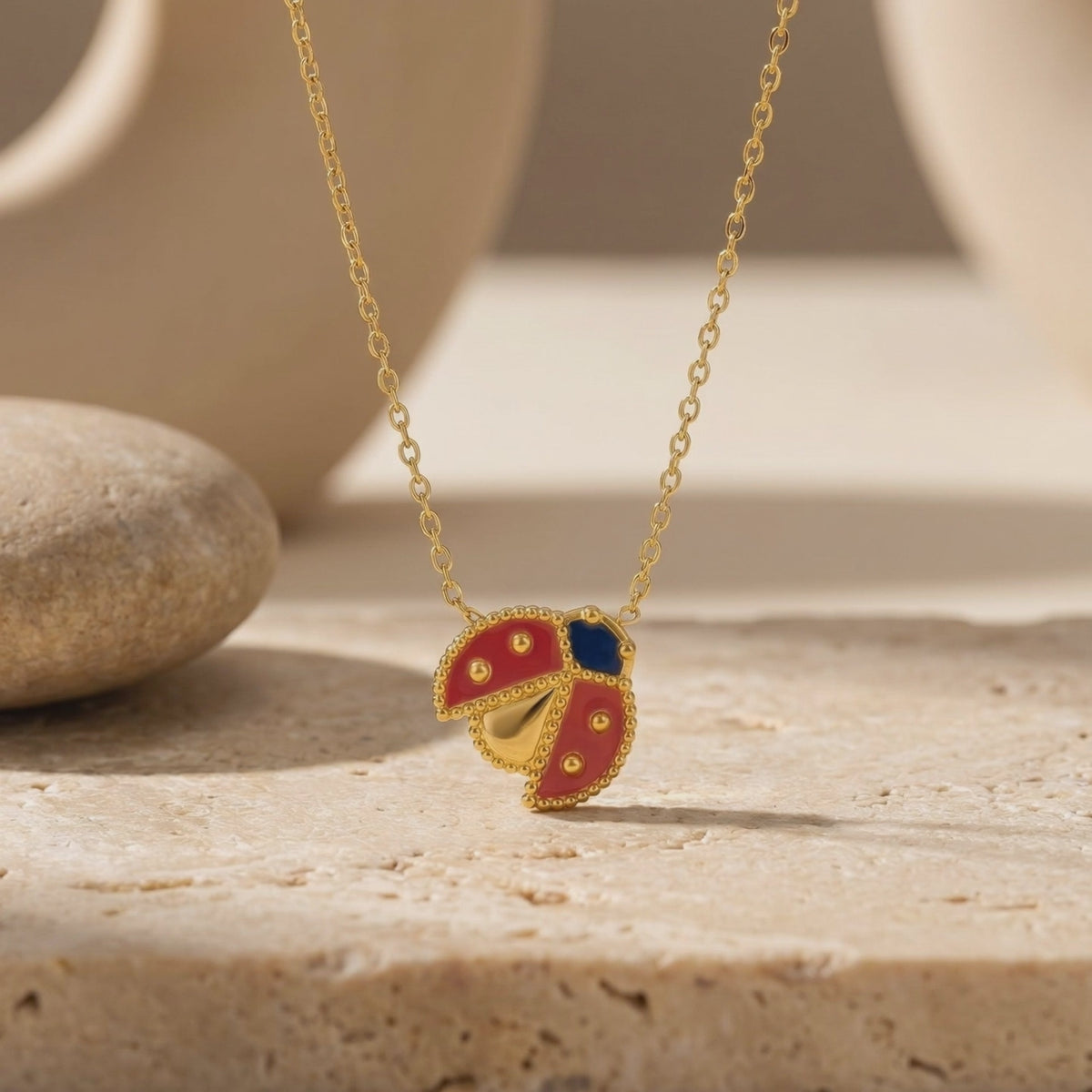 Gold ladybug pendant necklace with red and blue enamel detailing displayed on a textured stone surface.