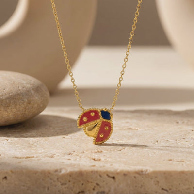 Gold ladybug pendant necklace with red and blue enamel detailing displayed on a textured stone surface.