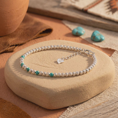 Pecos silver bead bracelet with turquoise accents and a heart charm displayed on a tan stone pedestal.