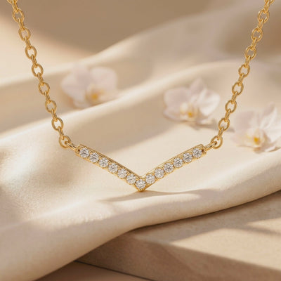 Close-up of a gold-tone chevron pendant with round clear stones on cream fabric, with white flowers in the background.