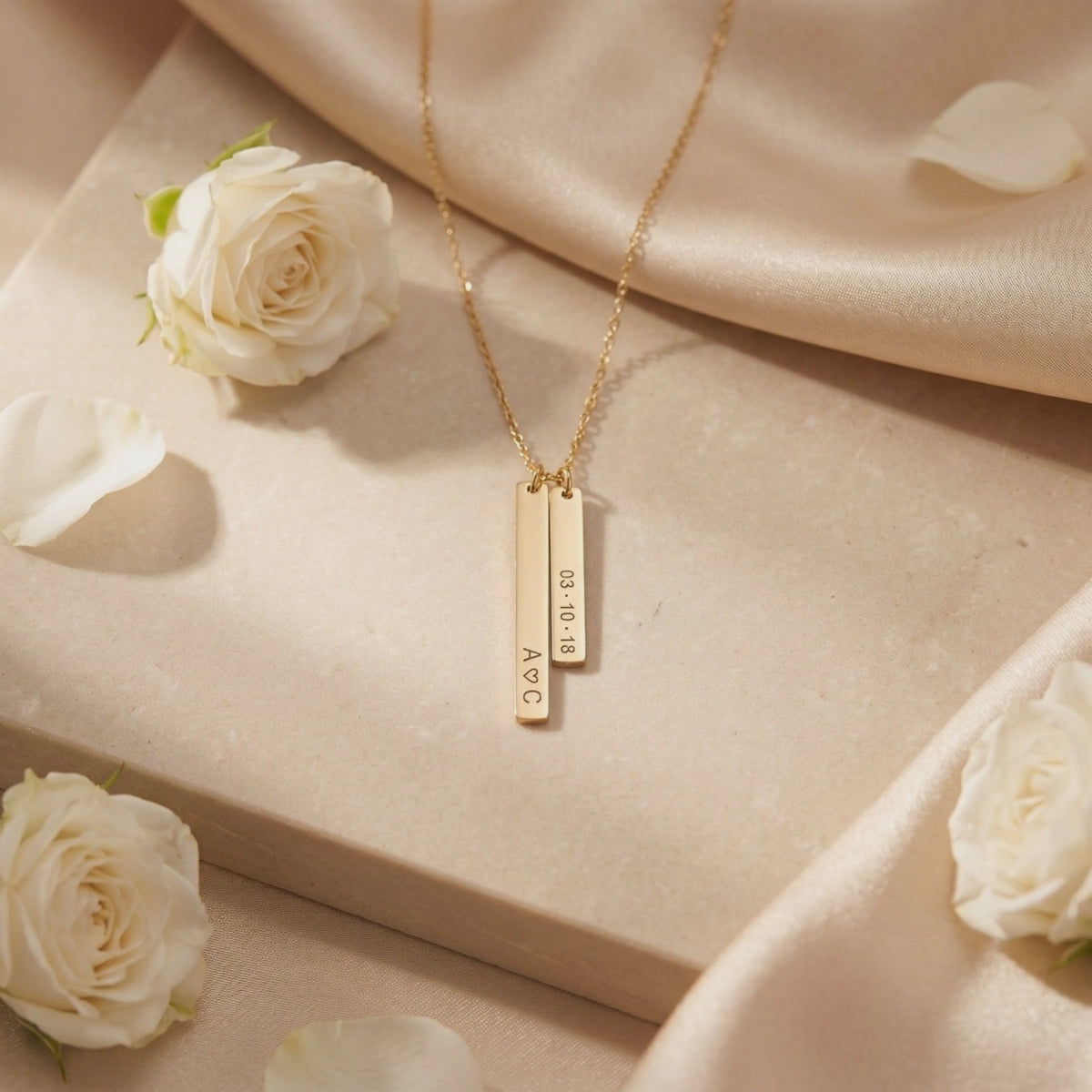 Flat lay of a gold-tone vertical bar necklace engraved with initials and a date, set among white roses.