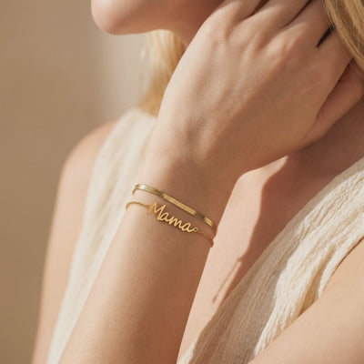 Gold-toned double-layer bracelet with flat and cable chains, worn on a wrist with a visible “Mama” centerpiece.