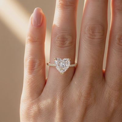 Top view of a gold-tone ring with a large heart-cut clear center stone and pavé band worn on a finger