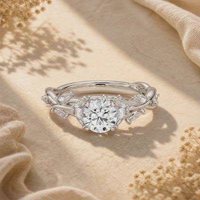 Front-facing close-up of the silver round moissanite ring displayed on beige fabric, showing leaf-and-vine side details.
