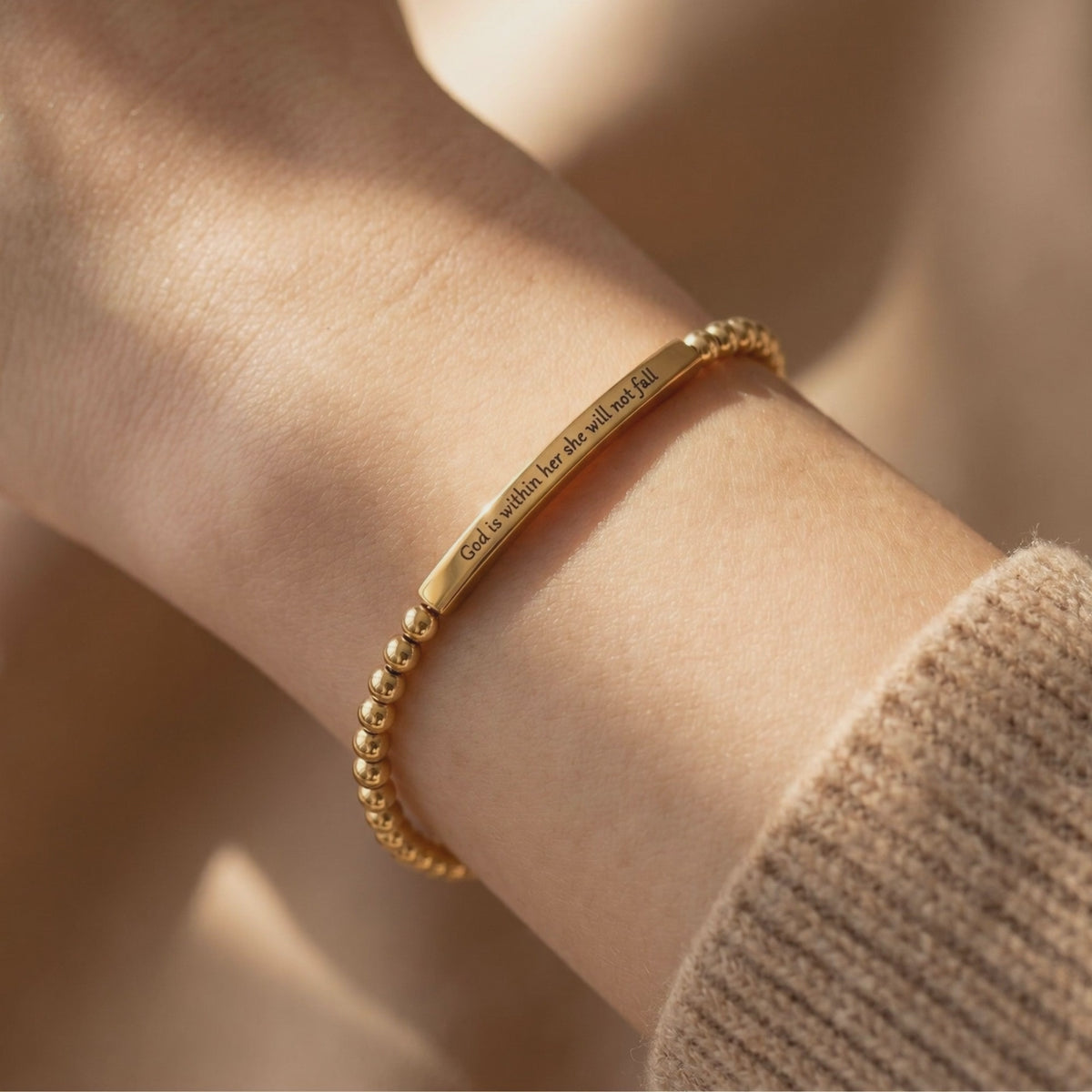 Gold-toned beaded bracelet with a bar engraved “God is within her she will not fall,” worn on a wrist.