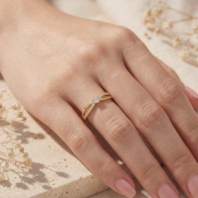 Gold-toned bypass ring with a round center stone and pavé accents, shown worn on a hand.