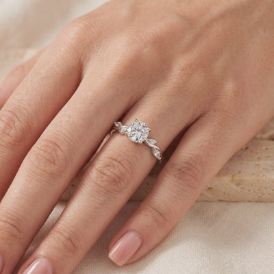 Silver leaf-motif ring with a round clear center stone worn on a hand over neutral fabric.