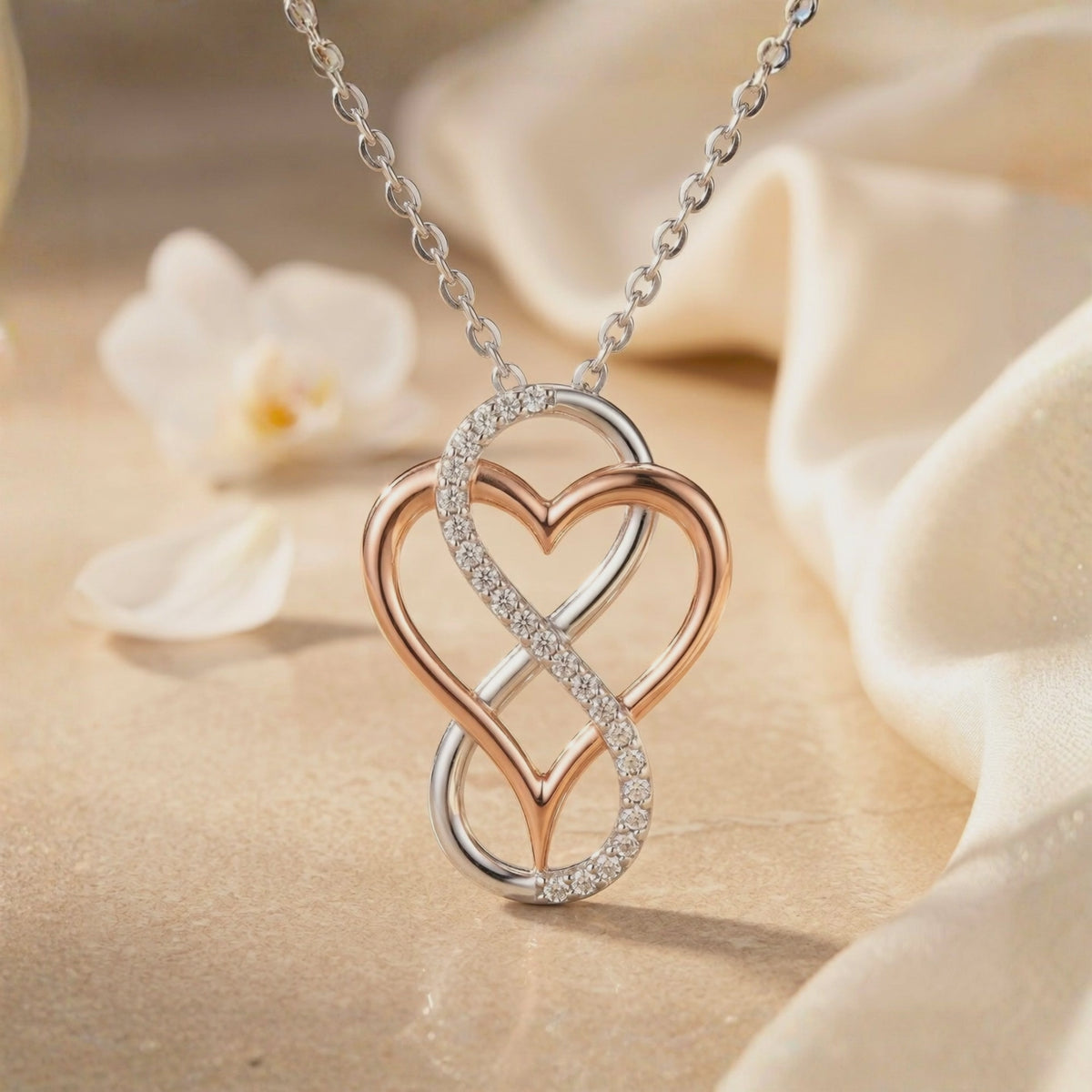 Close-up of a two-tone infinity-heart pendant with a pavé stone-accented loop, displayed beside white flowers and fabric.
