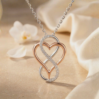 Close-up of a two-tone infinity-heart pendant with a pavé stone-accented loop, displayed beside white flowers and fabric.
